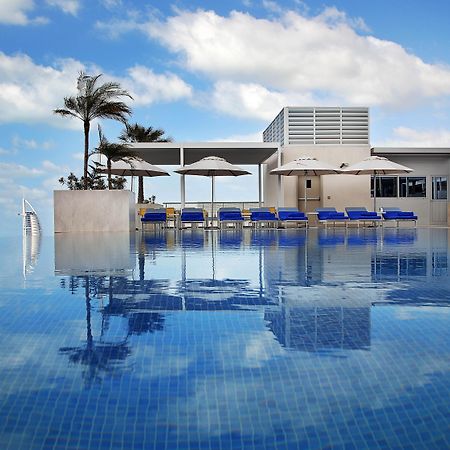 โรงแรมแกรนด์ คอสโมโพลิตัน - ดูไบ ภายนอก รูปภาพ The photo depicts a luxurious swimming pool with a stunning infinity edge. The water is calm and reflects the sky, creating a serene atmosphere. Surrounding the pool are several blue lounge chairs with white umbrellas, providing spots for relaxation.