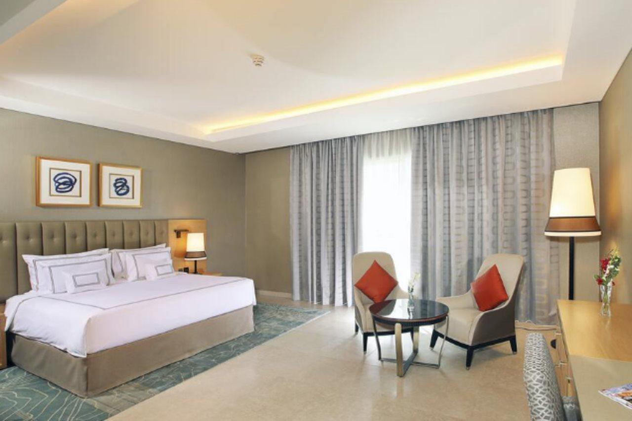 โรงแรมแกรนด์ คอสโมโพลิตัน - ดูไบ ภายนอก รูปภาพ The photo shows a modern hotel room featuring a spacious layout. There is a large bed with a headboard adorned with decorative artwork above it. On one side of the bed, there's a bedside lamp. Opposite the bed, there are two comfortable armchairs sty