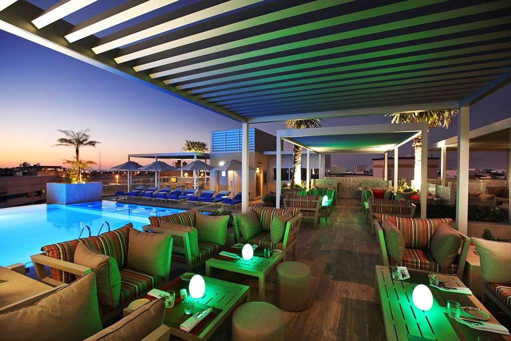 โรงแรมแกรนด์ คอสโมโพลิตัน - ดูไบ สิ่งอำนวยความสะดวก รูปภาพ The photo shows a stylish outdoor lounge area, likely at a hotel or resort. There are comfortable seating arrangements with striped cushions and small tables, illuminated with soft green lights. In the background, there is a swimming pool with lounge