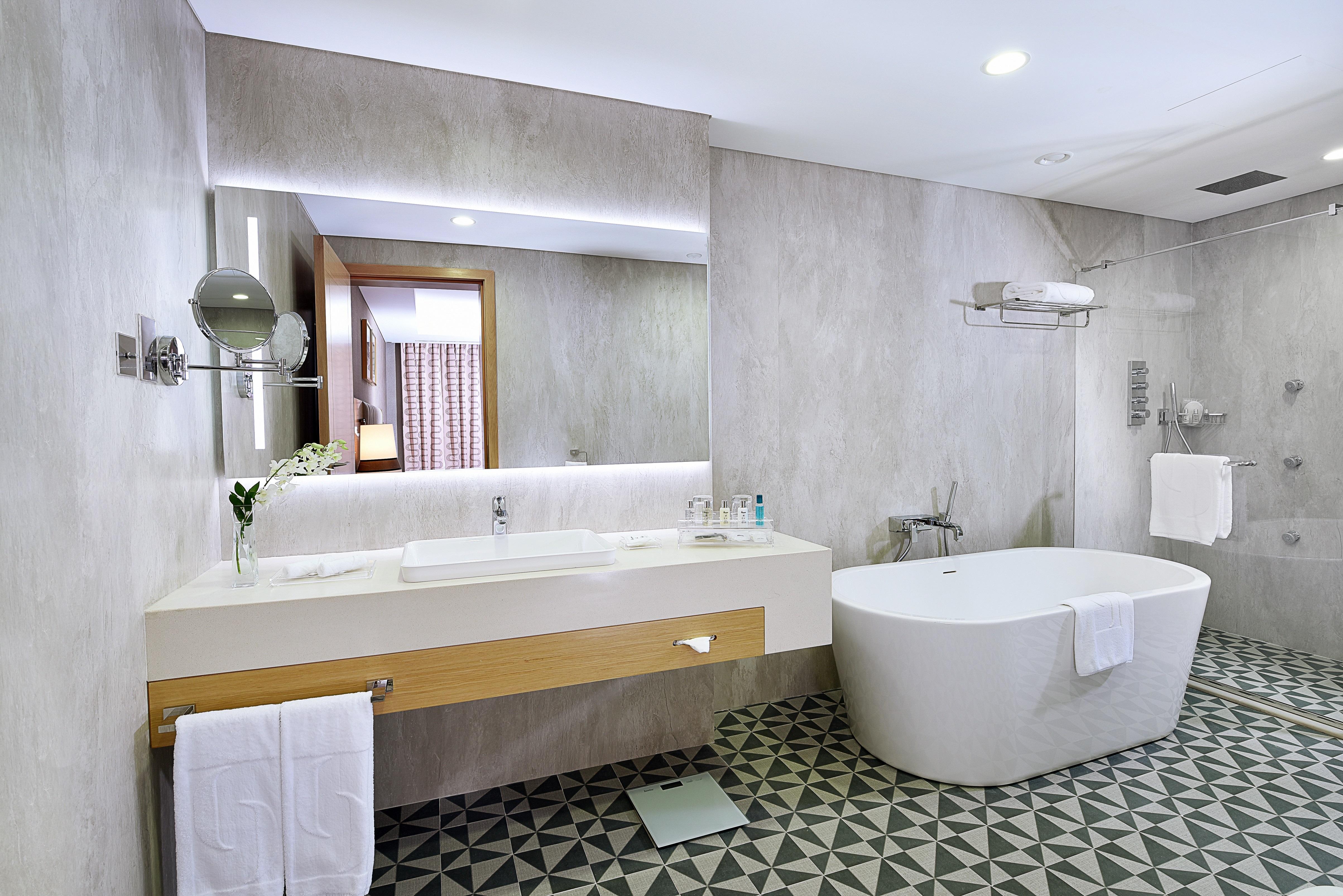 โรงแรมแกรนด์ คอสโมโพลิตัน - ดูไบ ภายนอก รูปภาพ The photo shows a modern bathroom featuring a sleek design. There is a large white freestanding bathtub on one side. The vanity area has a long countertop with a sink and a large mirror above it, illuminated by soft lighting. The walls are a light gr
