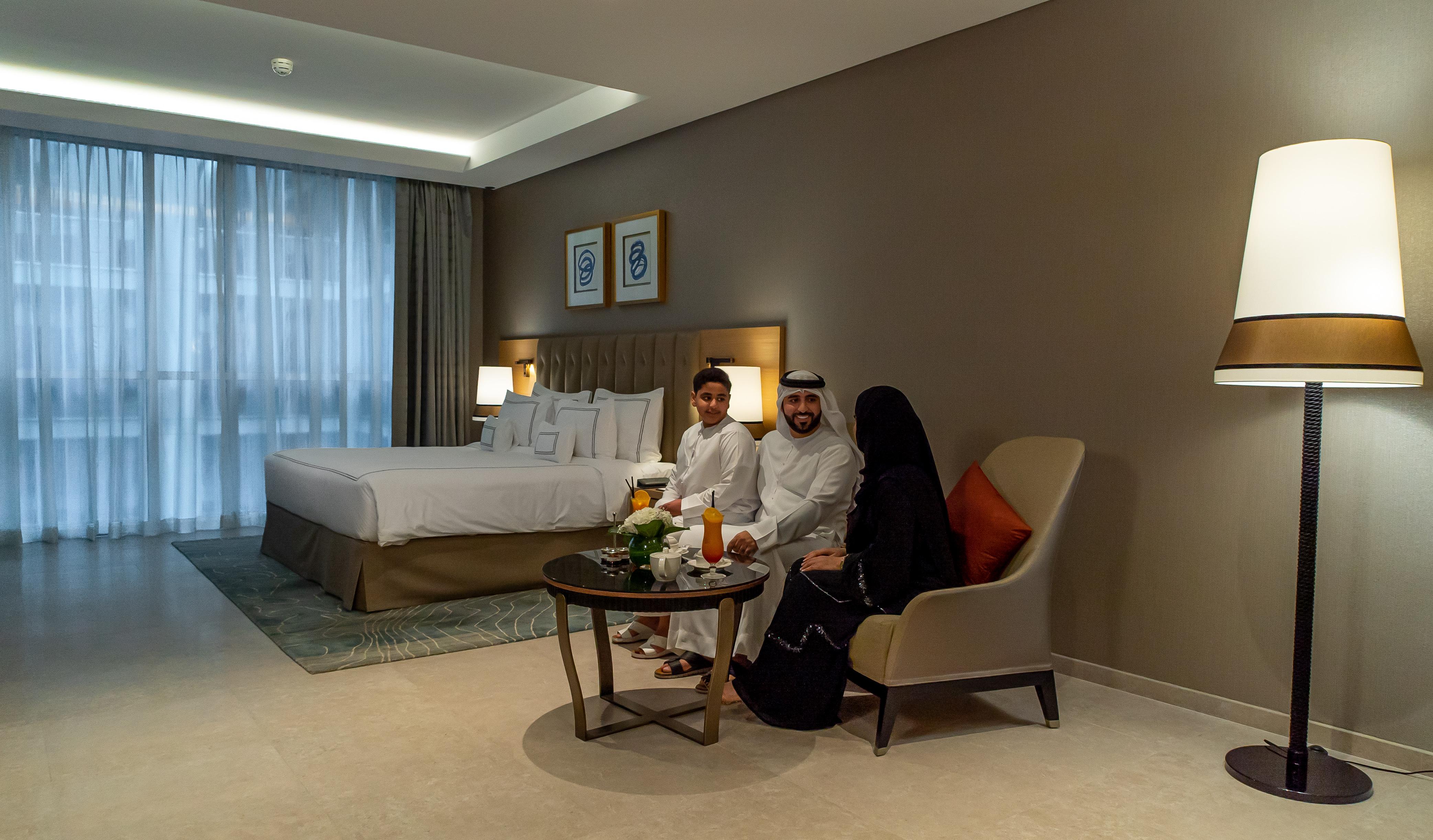 โรงแรมแกรนด์ คอสโมโพลิตัน - ดูไบ ภายนอก รูปภาพ The photo shows a modern hotel room featuring a large bed with white linens and a stylish headboard. In the room, there is a small round table with a floral centerpiece and drinks. Three individuals are seated on the chairs by the table: two adults a