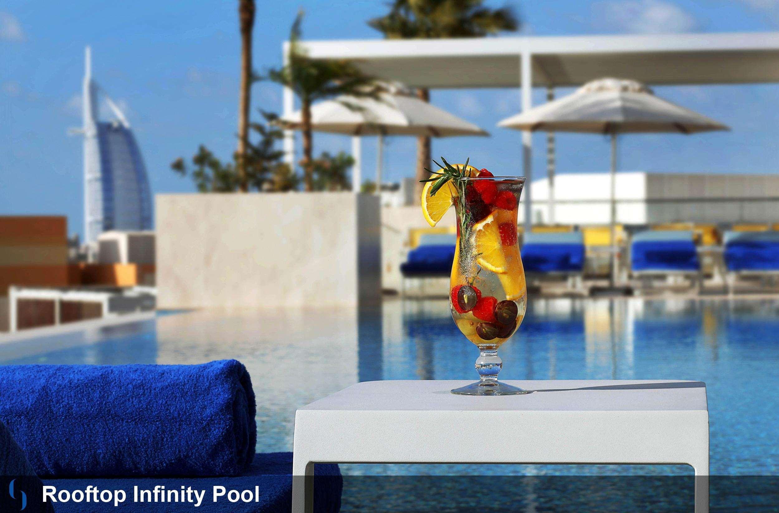โรงแรมแกรนด์ คอสโมโพลิตัน - ดูไบ ภายนอก รูปภาพ The photo shows a rooftop infinity pool with a stunning view. In the foreground, there is a cocktail glass filled with a colorful drink garnished with fruits, including lemon and berries. The background features cozy loungers and umbrellas, suggestin