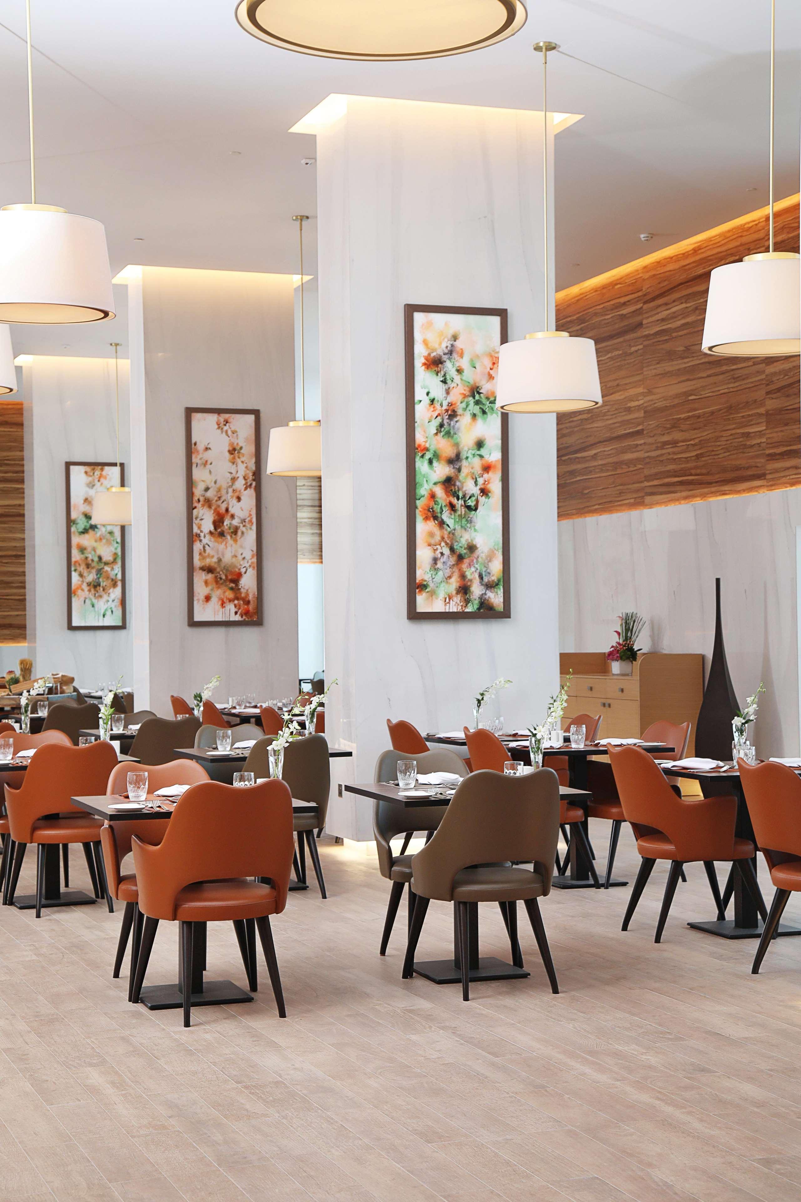 โรงแรมแกรนด์ คอสโมโพลิตัน - ดูไบ ภายนอก รูปภาพ The photo shows the interior of a modern restaurant. The space features elegantly set dining tables with glasses and cutlery, and the chairs have a mix of colors, including orange and neutral tones. There are large pieces of artwork on the walls, lik