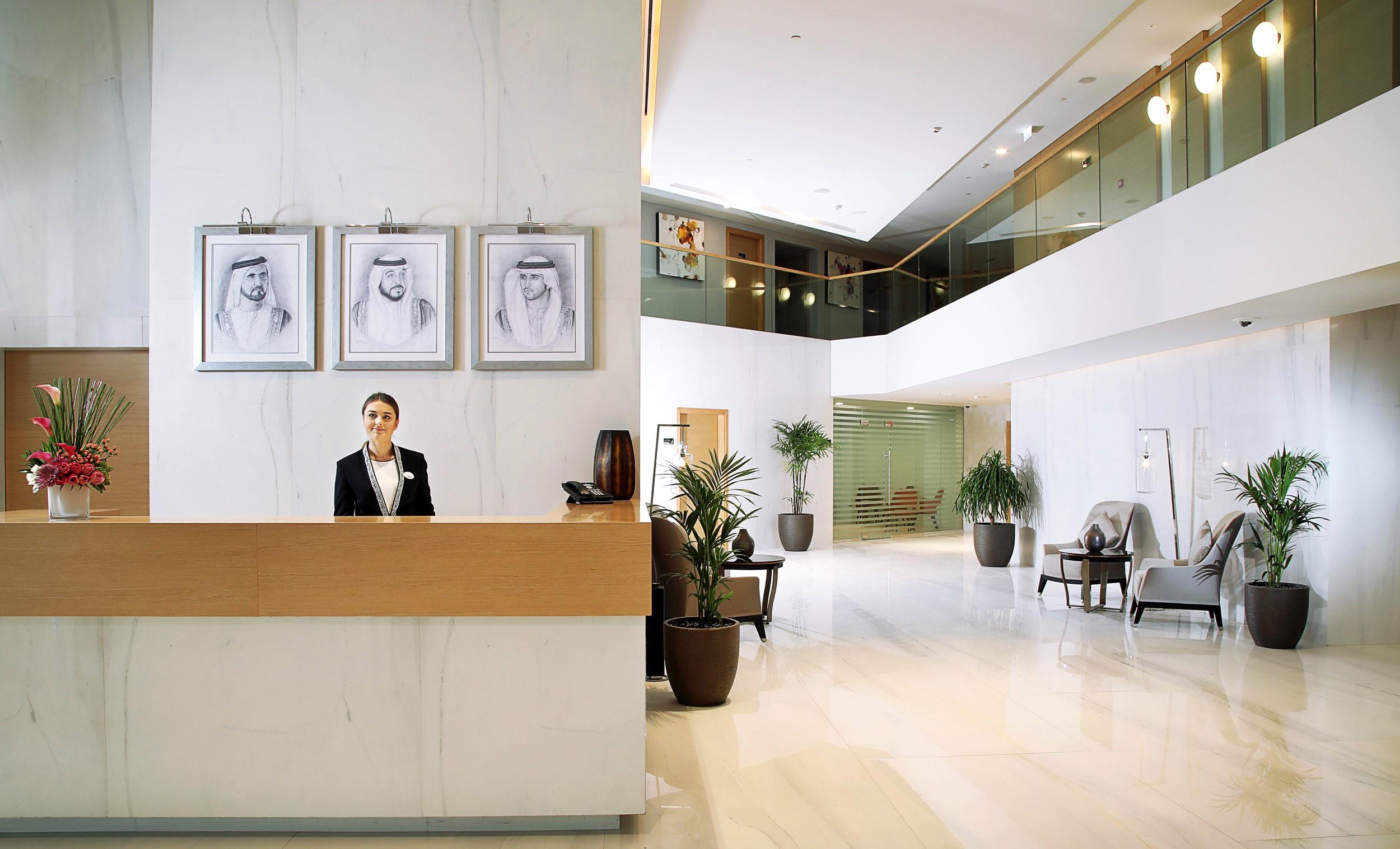 โรงแรมแกรนด์ คอสโมโพลิตัน - ดูไบ ภายนอก รูปภาพ The photo depicts a modern hotel lobby. In the foreground, there is a reception desk with a staff member dressed in formal attire, likely a receptionist. Behind the reception desk, three framed portraits are hanging on the wall, featuring individuals