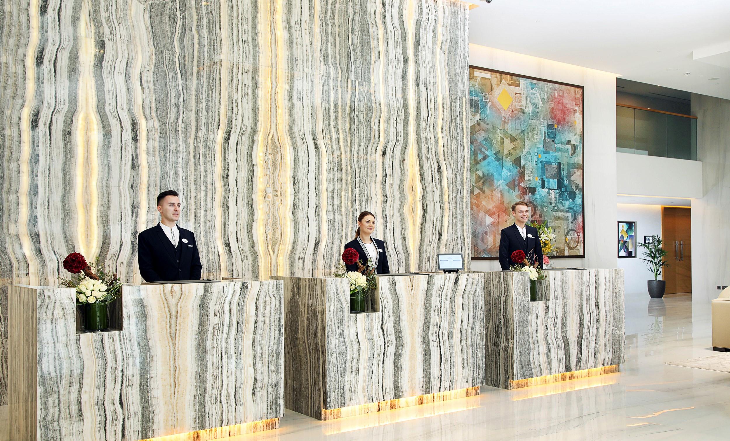 โรงแรมแกรนด์ คอสโมโพลิตัน - ดูไบ ภายนอก รูปภาพ The photo shows a hotel reception area featuring three receptionists standing behind a sleek, modern front desk made of a striped marble-like material with illuminated edges. They are dressed in formal uniforms. Behind them, there is a large piece of