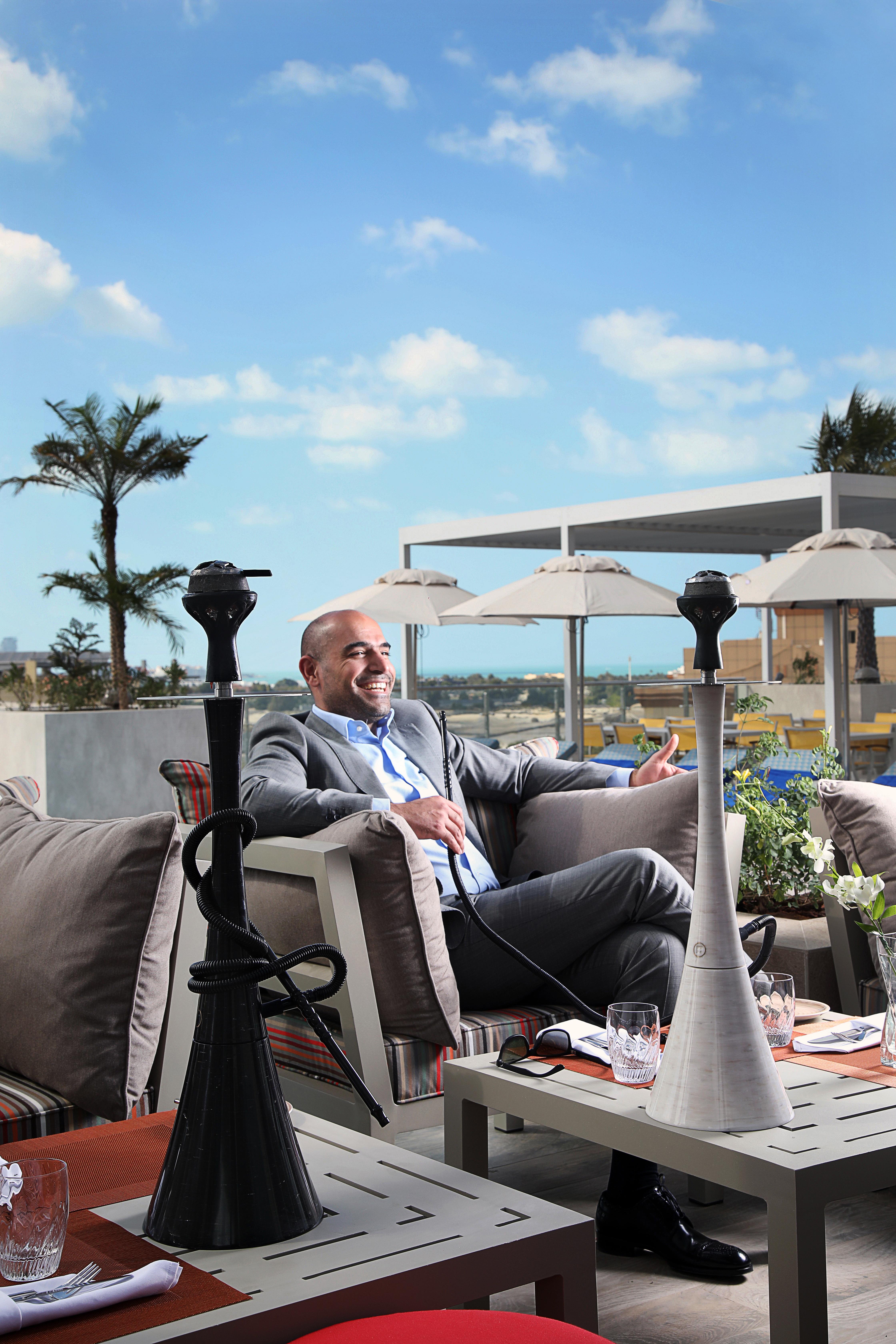 โรงแรมแกรนด์ คอสโมโพลิตัน - ดูไบ ภายนอก รูปภาพ The image shows a man sitting comfortably on a sofa in an outdoor lounge area. He is dressed in a suit and is smiling, suggesting a relaxed atmosphere. In front of him are two hookahs, which are traditional smoking devices. The background features pa