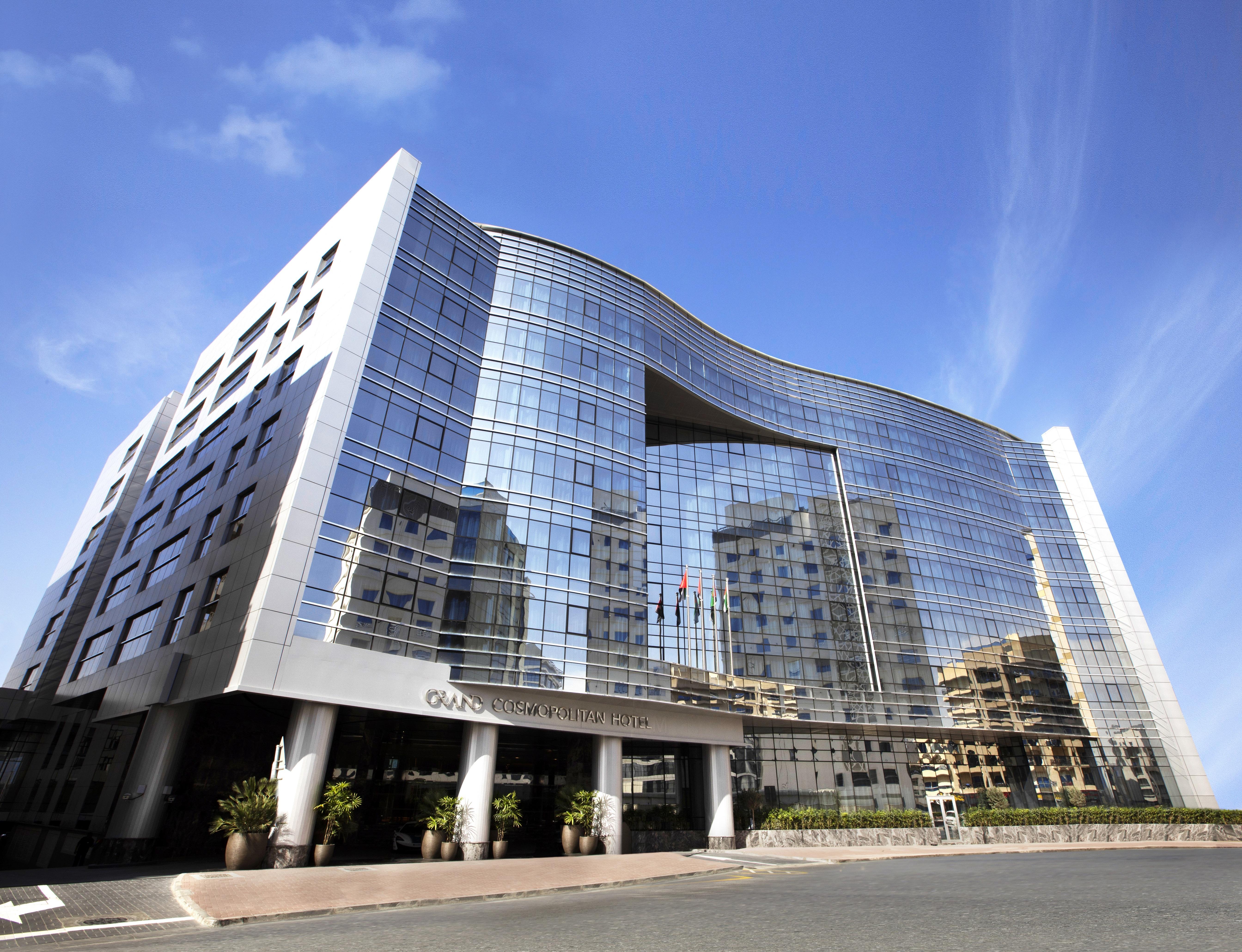 โรงแรมแกรนด์ คอสโมโพลิตัน - ดูไบ ภายนอก รูปภาพ The image shows a modern building with a distinctive architectural design. The facade is predominantly made of glass, reflecting the blue sky and surrounding structures. The building has a curved top, creating a unique silhouette, and is likely multi