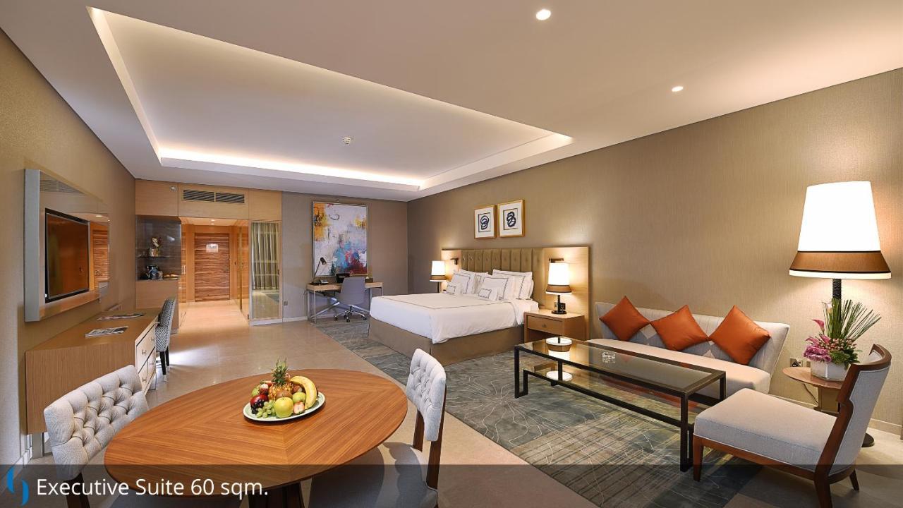 โรงแรมแกรนด์ คอสโมโพลิตัน - ดูไบ ภายนอก รูปภาพ The image shows a spacious hotel suite. It features a king-sized bed with a modern design, accompanied by decorative pillows. To one side, there is a seating area with a sofa and a glass coffee table. A dining table with chairs is also visible, posit