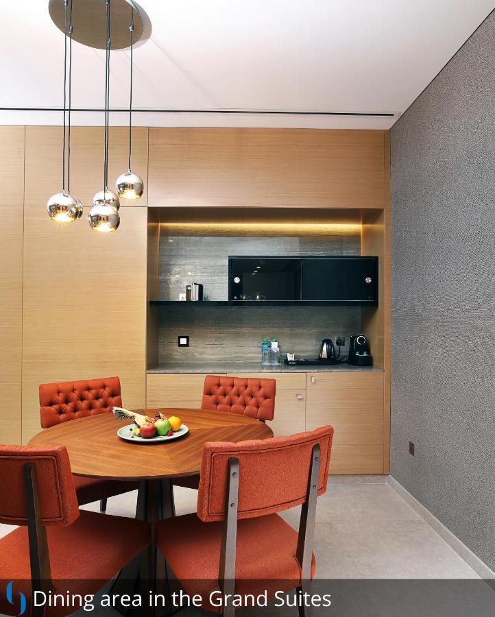 โรงแรมแกรนด์ คอสโมโพลิตัน - ดูไบ ภายนอก รูปภาพ The photo depicts a modern kitchen area. There is a round dining table with orange upholstered chairs surrounding it. On the table, a bowl of assorted fruits is displayed. In the background, a countertop with some kitchen appliances, including a kett