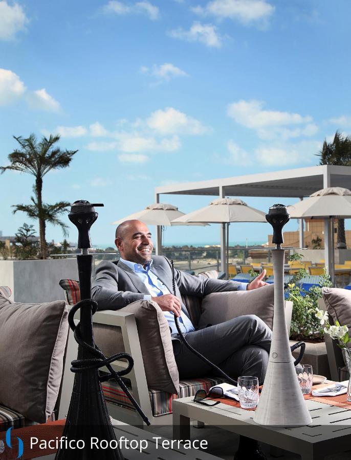 โรงแรมแกรนด์ คอสโมโพลิตัน - ดูไบ ภายนอก รูปภาพ The photo shows a man sitting comfortably in an outdoor lounge area. He is dressed in a light grey suit and appears to be smiling and relaxed. In the background, there are several palm trees and sun loungers with umbrellas, suggesting a tropical or u