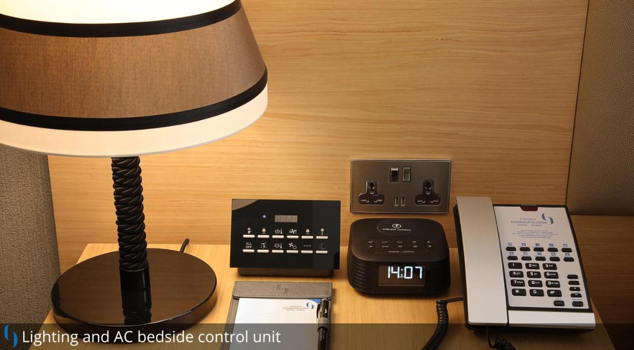 โรงแรมแกรนด์ คอสโมโพลิตัน - ดูไบ ภายนอก รูปภาพ The photo shows a bedside setup with several items. There is a table lamp with a dark lampshade on the left. Next to it is a control unit with various buttons, likely for managing room settings such as lighting and air conditioning. Beside the contro