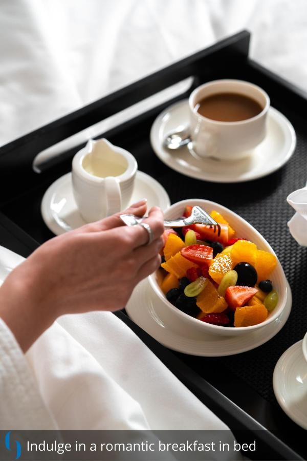 โรงแรมแกรนด์ คอสโมโพลิตัน - ดูไบ ภายนอก รูปภาพ The photo displays a hand holding a fork, poised to pick up fruit from a bowl. The bowl contains a colorful assortment of fresh fruit, including pieces of orange, strawberries, grapes, and blueberries. Nearby, there is a cup of coffee and a small sau