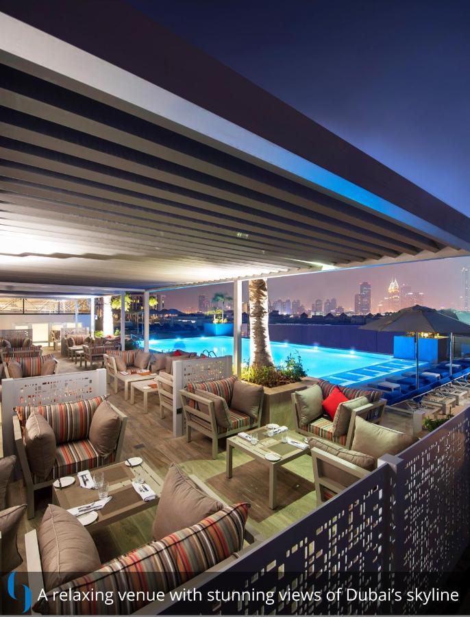 โรงแรมแกรนด์ คอสโมโพลิตัน - ดูไบ ภายนอก รูปภาพ The photo depicts a stylish outdoor lounge area at night. It features a modern design with comfortable seating arrangements, including plush sofas and tables under a canopy. In the background, there is a swimming pool illuminated with blue lights, cr