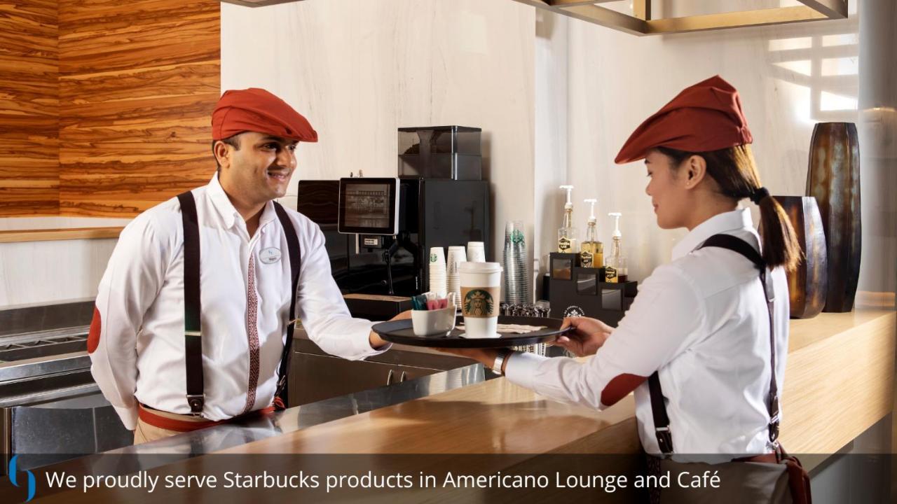 โรงแรมแกรนด์ คอสโมโพลิตัน - ดูไบ ภายนอก รูปภาพ The photo shows two staff members in a café environment. One is handing a tray with a Starbucks beverage to the other, who is standing at the counter. Both employees are wearing white shirts with suspenders and red caps. The background suggests a mod