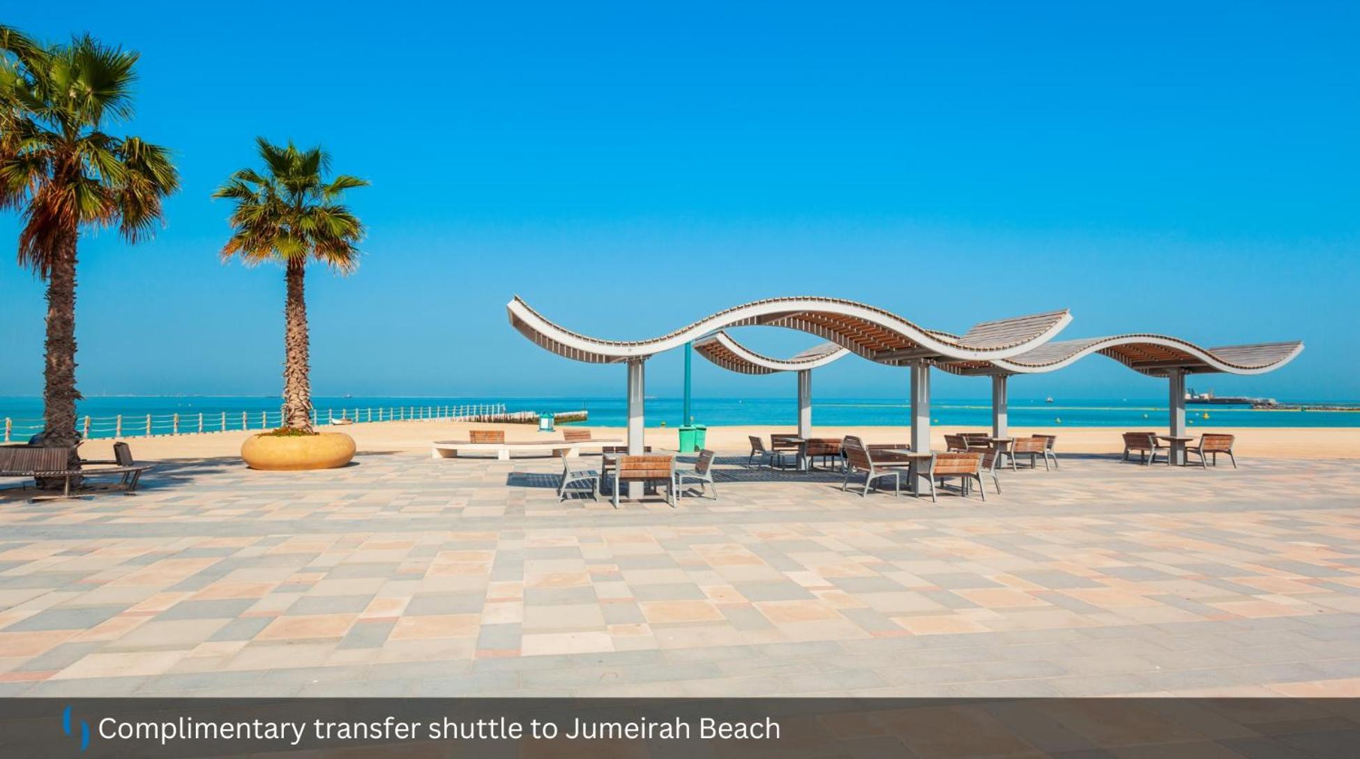 โรงแรมแกรนด์ คอสโมโพลิตัน - ดูไบ ภายนอก รูปภาพ The photo shows a scenic beach area, likely at Jumeirah Beach. In the foreground, there are wooden benches and tables arranged under shaded structures with a wavy design. A tall palm tree adds to the tropical feel of the setting. In the background, y