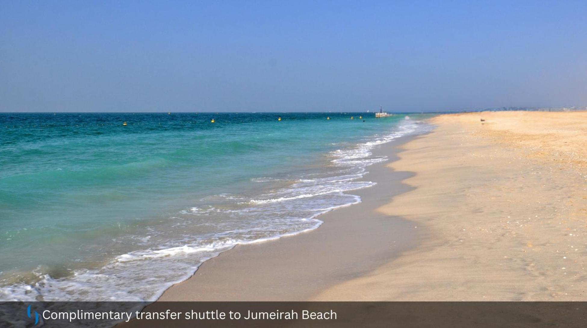 โรงแรมแกรนด์ คอสโมโพลิตัน - ดูไบ ภายนอก รูปภาพ The photo depicts a serene beach scene featuring soft, golden sand and clear turquoise waters. Gentle waves are lapping at the shore, creating a peaceful ambiance. The sky above is mostly clear, suggesting a bright and sunny day. There are some buoys